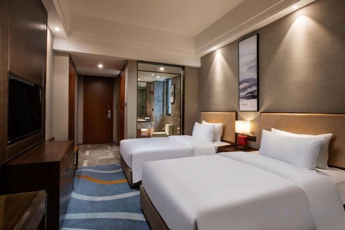 A hotel room with two beds, a TV, a desk, and a bathroom visible through an open door.