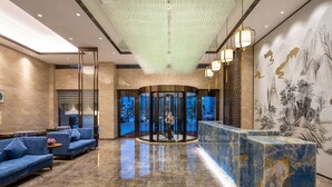 A lobby with a marble reception desk, blue sofas, a large chandelier, and a mural on the wall. - Ramada Encore by Wyndham Wuhan Caidian (Wuhan)