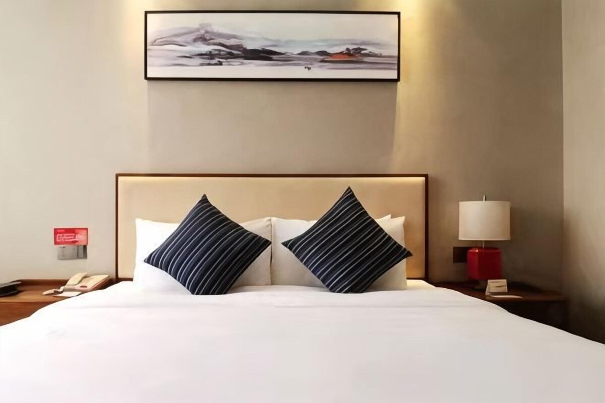 Habitación de hotel con cama, dos almohadas, mesita de noche con lámpara, teléfono y un cuadro en la pared.