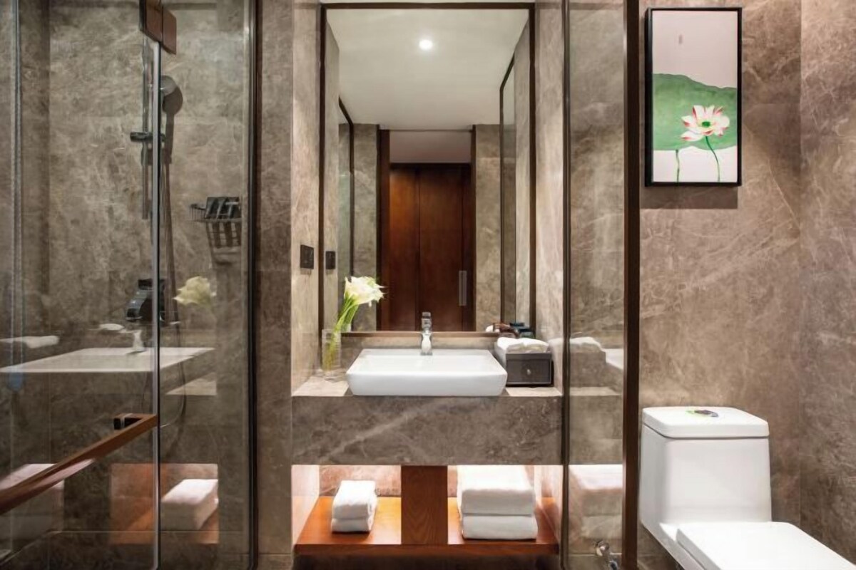 Un baño moderno con ducha de vidrio, un lavamanos central con encimera de mármol y un mueble de tocador de madera con dos toalleros.