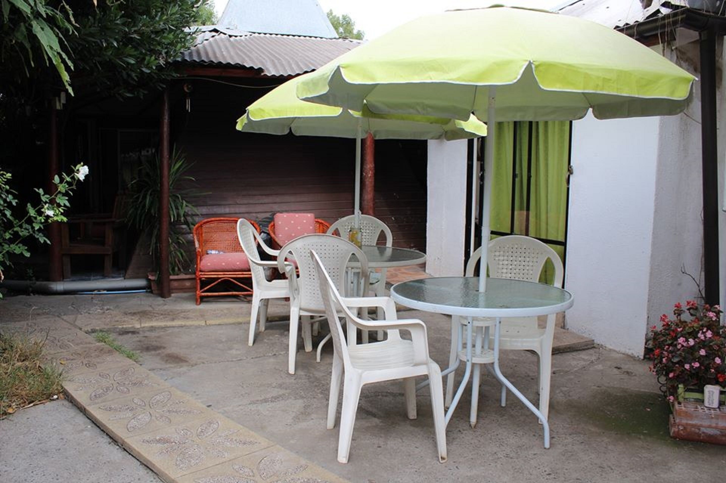 An outdoor patio with a round glass table and white plastic chairs, shaded by a green umbrella.