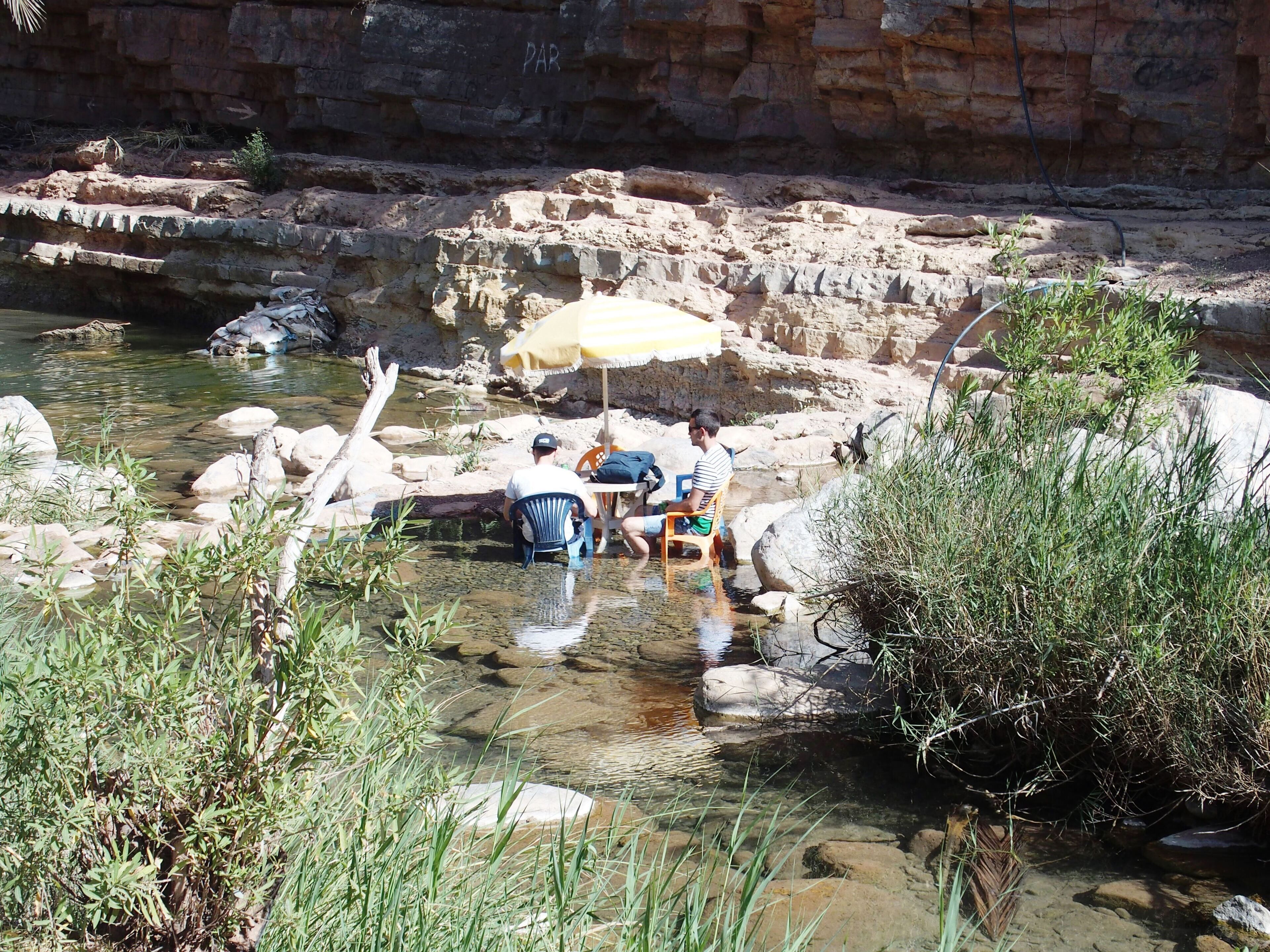 Un cadre naturel avec une rive rocheuse, une eau claire et quelques personnes assises au bord de l’eau.