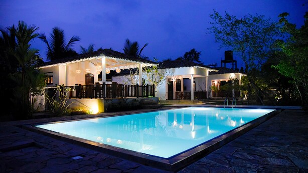 Vista nocturna de un área de piscina con una casa al fondo, rodeado de palmeras y un camino de piedra.