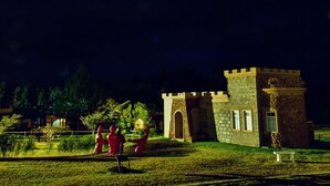 A stone building with arched windows and a small tower, surrounded by a well-lit garden with statues and a pond. - Tafaria Castle (Ndaragwa)