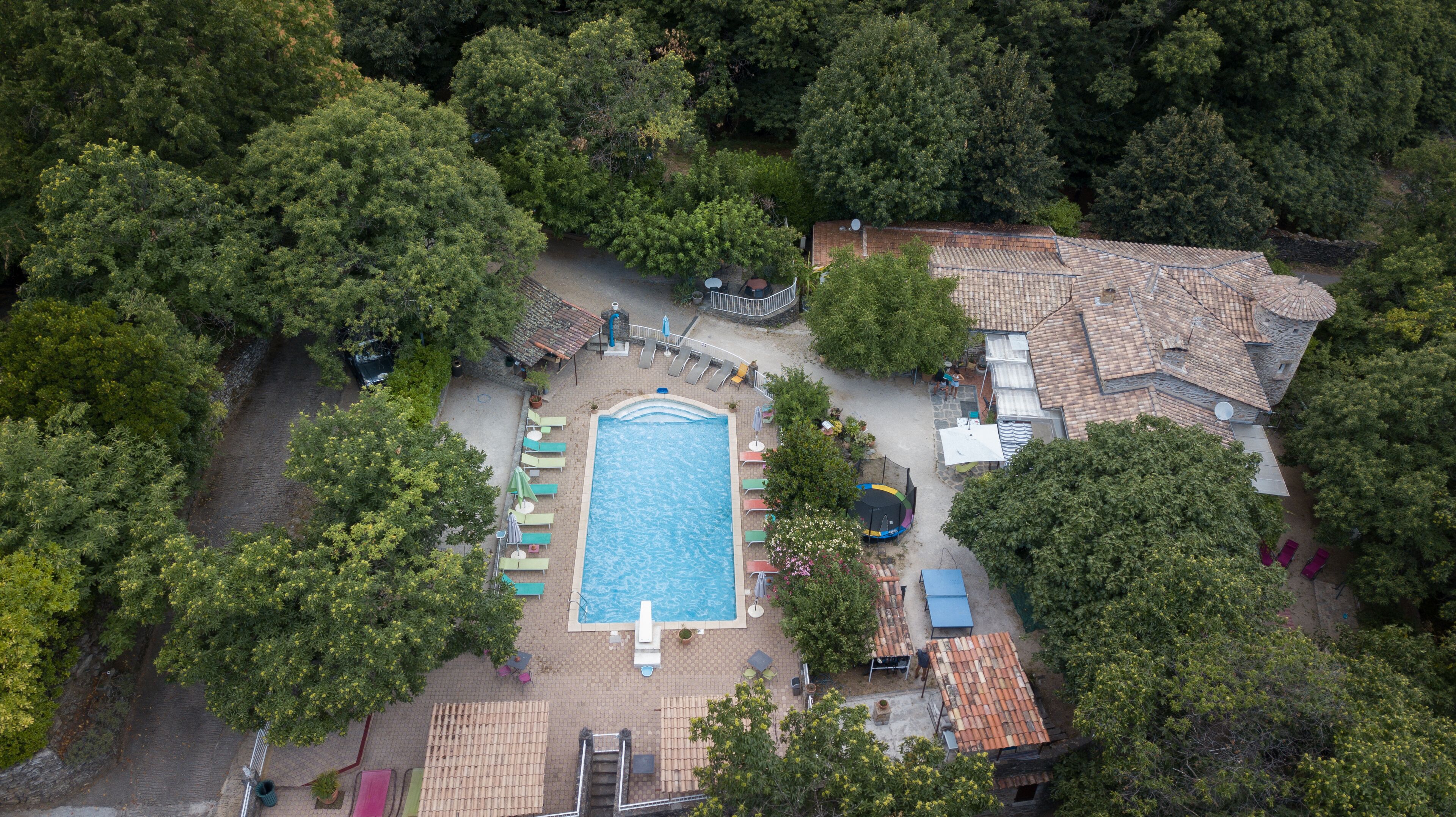 An aerial view of a property with a swimming pool surrounded by lounge chairs and umbrellas, a stone building with a tower, and dense surrounding trees.
