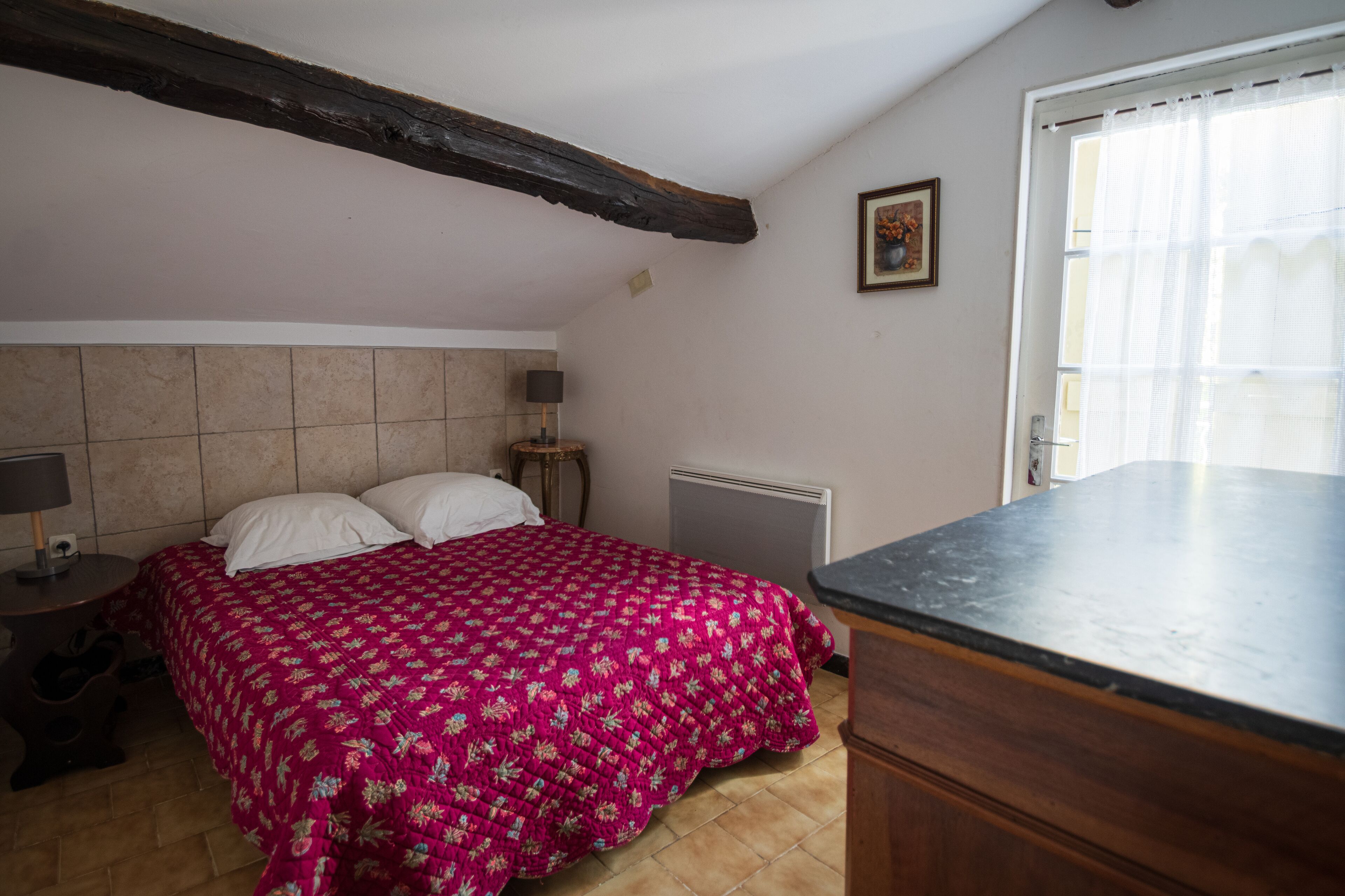 Une chambre à coucher comprenant un lit, une table de chevet, une lampe, un radiateur, une fenêtre avec des rideaux et une poutre en bois au plafond.
