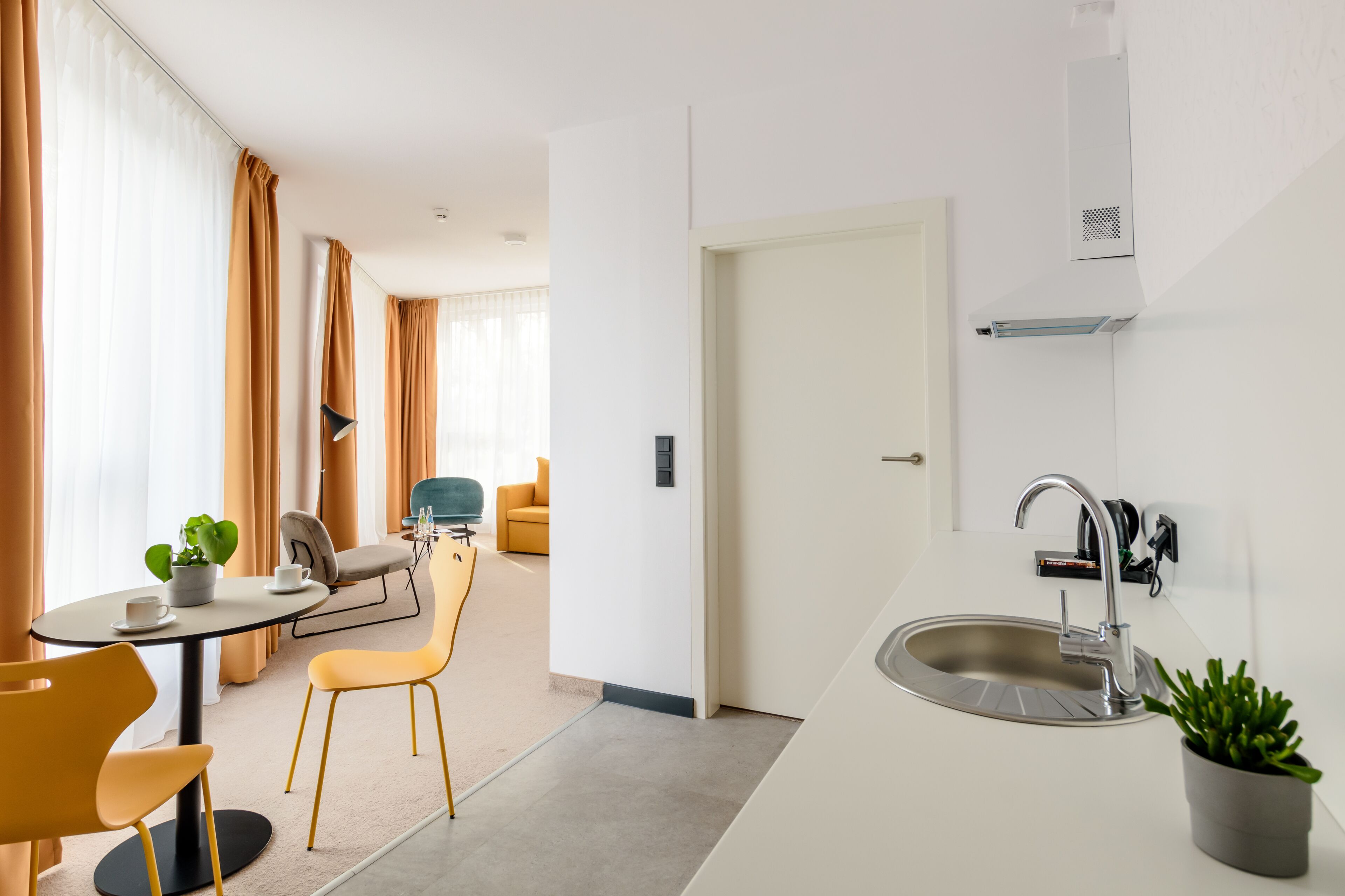 A modern kitchen with a sink, a dining area with yellow chairs, and a view into a living room with a sofa and curtains.