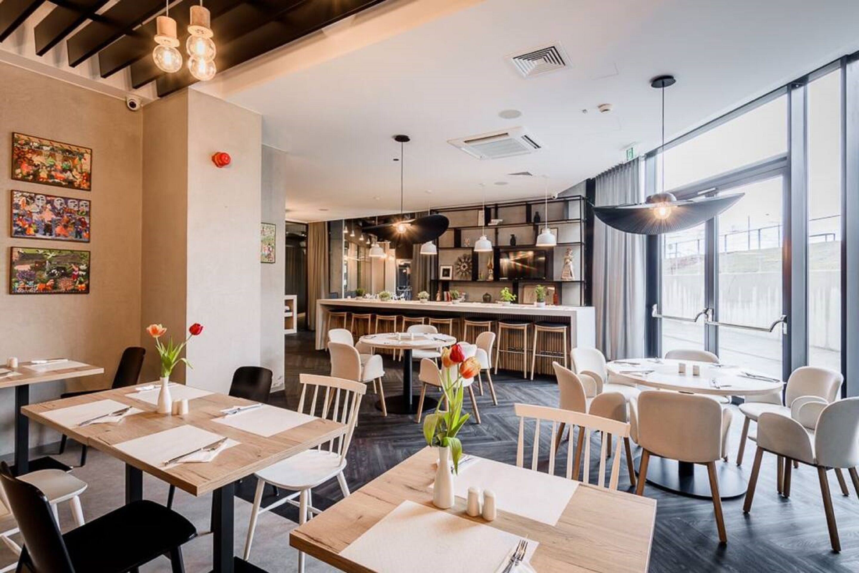 A modern restaurant with wooden tables and chairs, a central bar area, and large windows providing natural light.