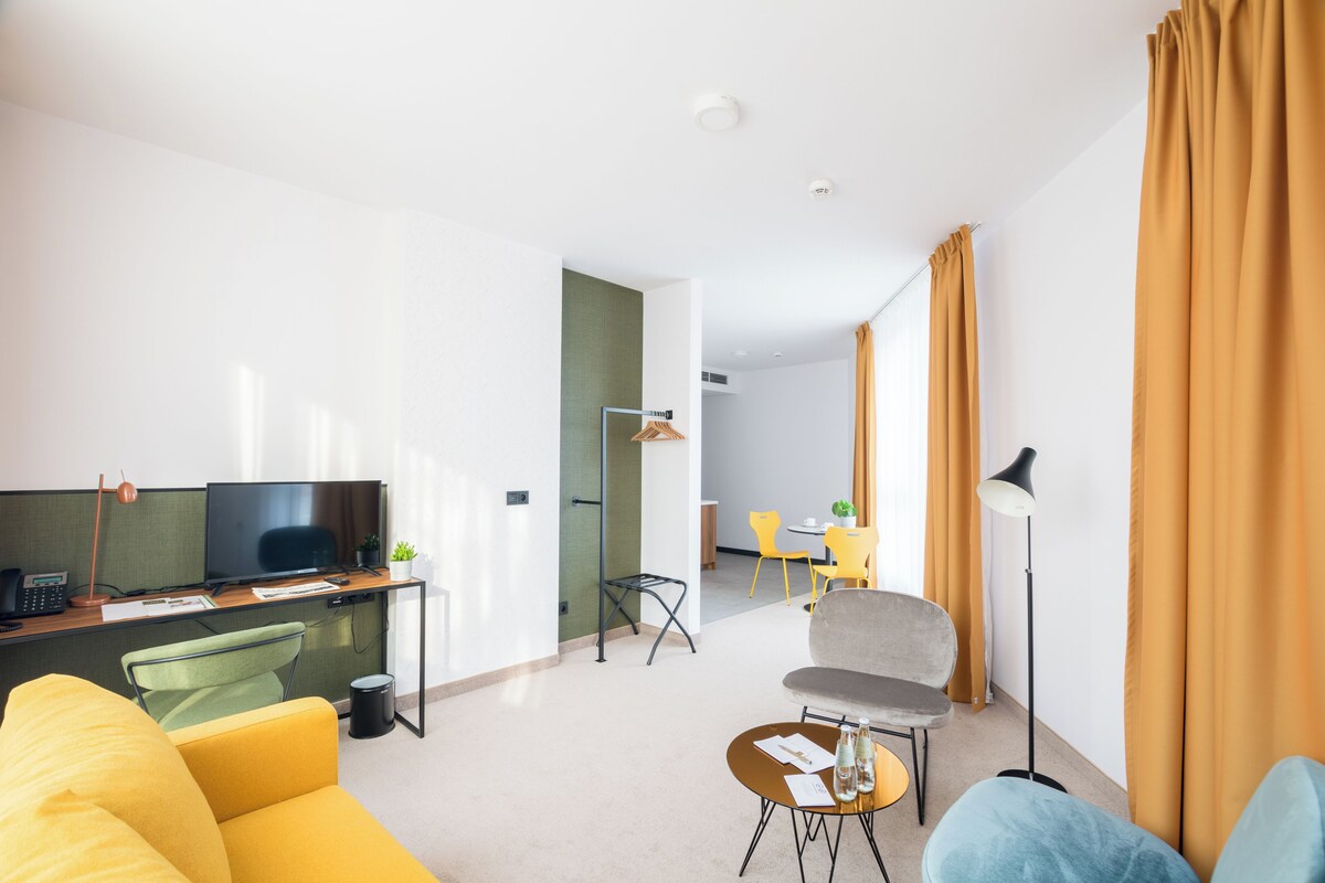 A modern living room with a sofa, a coffee table, a TV, and a desk. There is a dining area in the background.