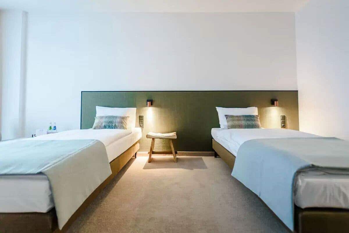 A hotel room with two single beds, a small stool, and a nightstand with a lamp.