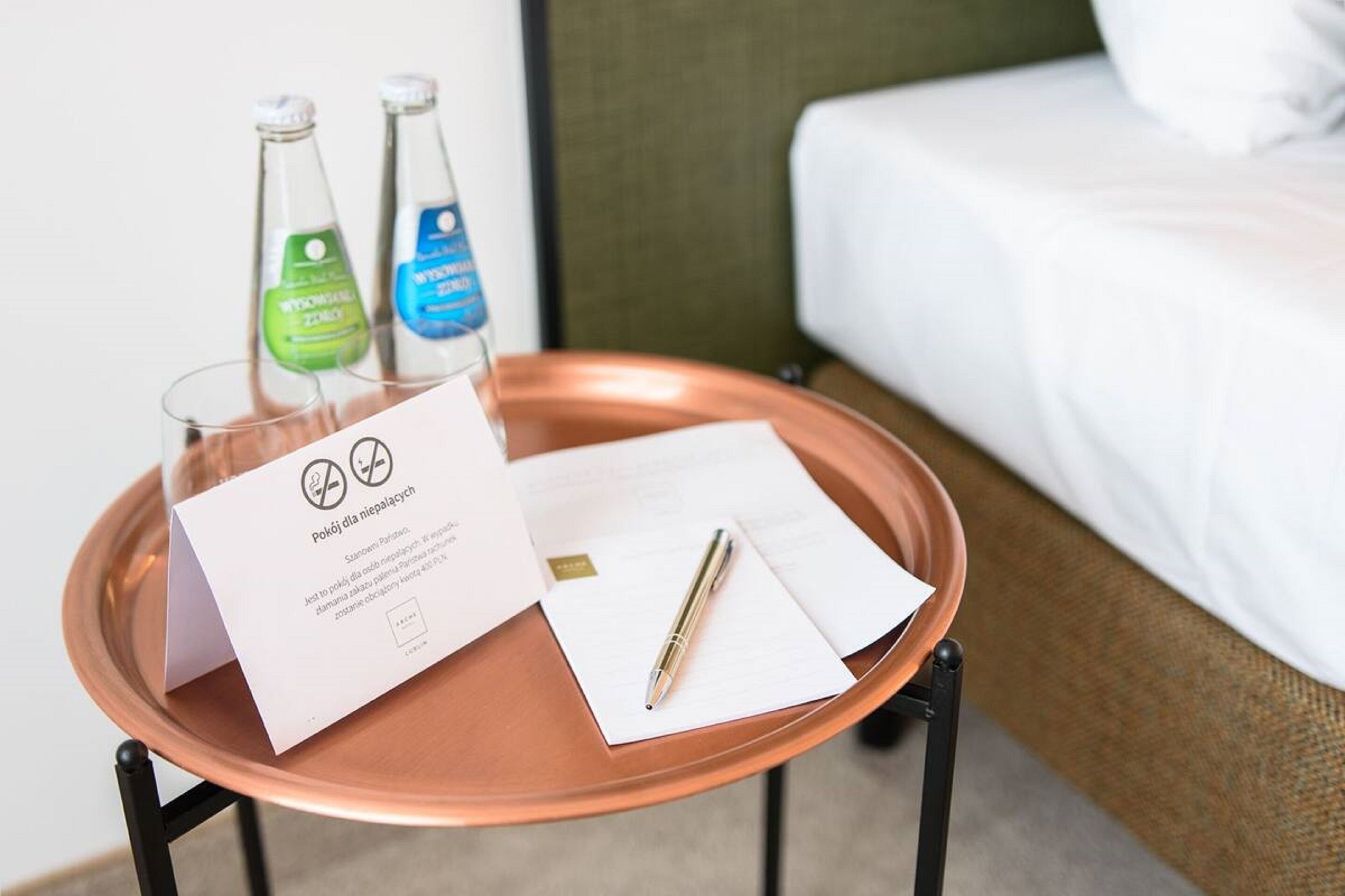 A hotel room with a bed, a small table with a tray holding a glass, two bottles, a pen, and a card.