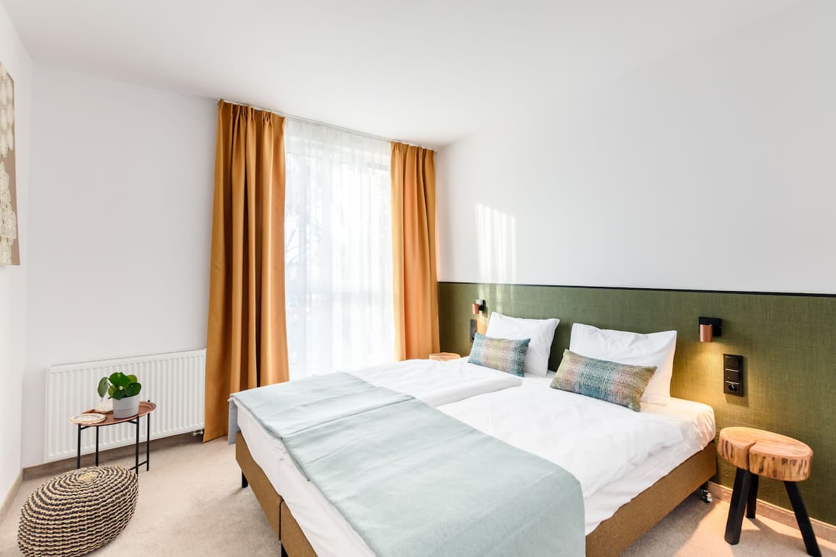 A modern hotel room with a large bed, a bedside table, a small stool, a radiator, and a window with curtains.