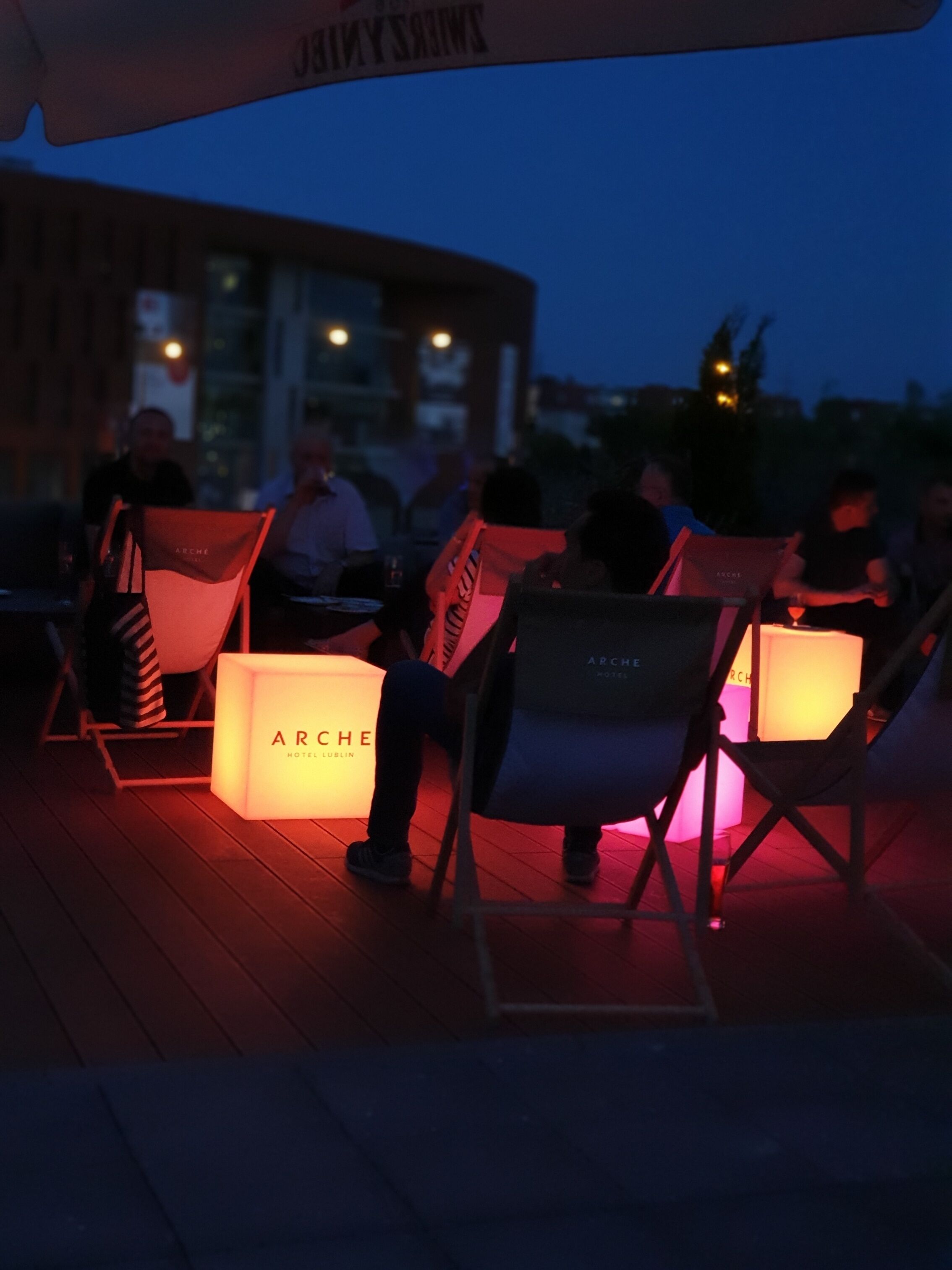 Terrasse mit beleuchteten Tischen und Stühlen, ein Holzdeck und ein Gebäude im Hintergrund.