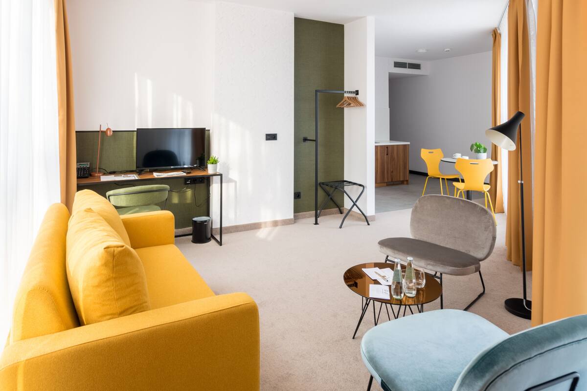 A modern hotel room with a yellow sofa, a blue armchair, a small round table with a lamp, a TV, and a desk with a computer and a plant.