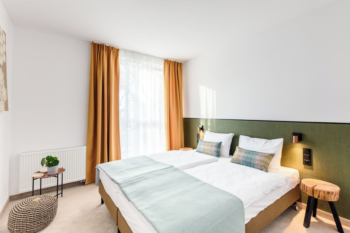Habitación de hotel moderna con una cama grande, una mesita de noche, un taburete pequeño, un radiador y una ventana con cortinas.
