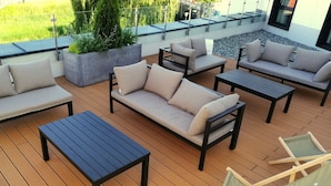 Terraza con mobiliario exterior, incluyendo sofás, una mesa y sillas, rodeada de vegetación y barandilla de cristal.