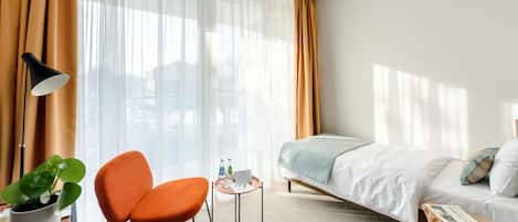 Una habitación de hotel con una cama, una silla naranja, una mesita con una lámpara y una planta.