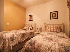 Een kamer met twee eenpersoonsbedden, elk voorzien van een gewatteerde spreidek en bijpassende kussens. Twee ingelijste foto’s hangen aan de muur, een nachtkastje met een lamp en een klein decoratief object op het nachtkastje.