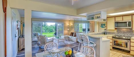 Una cocina amplia con electrodomésticos modernos, un comedor con mobiliario de mimbre y ventanas grandes con vistas al exterior.