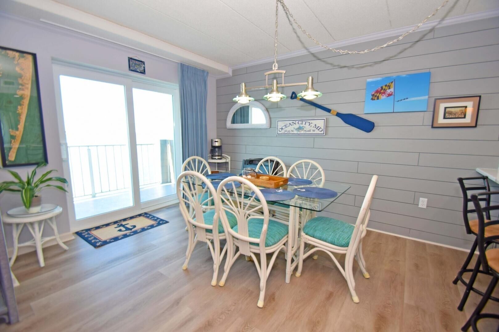 A dining area with a glass table and white chairs, a blue and white nautical-themed rug, and a view of the ocean through a sliding door.