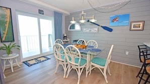 Un comedor con mesa de vidrio y sillas blancas, un tapete azul y blanco con motivos náuticos, y vista al océano a través de una puerta corrediza.