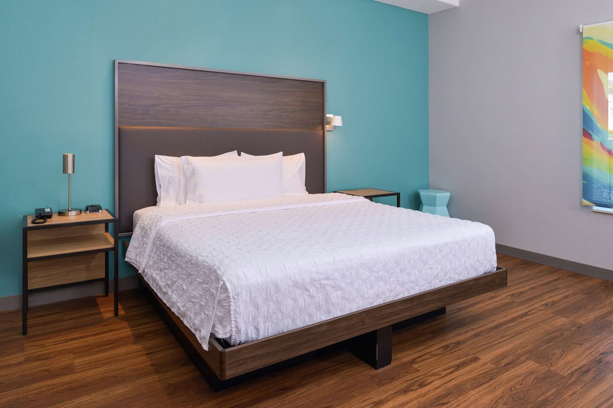 Una habitación de hotel moderna con un marco de cama de madera, una colcha blanca, una mesita de noche con una lámpara y una pequeña mesita auxiliar.