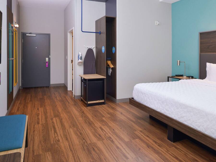 Habitación de hotel con piso de madera, cama con ropa blanca, escritorio con silla, armario y espejo.
