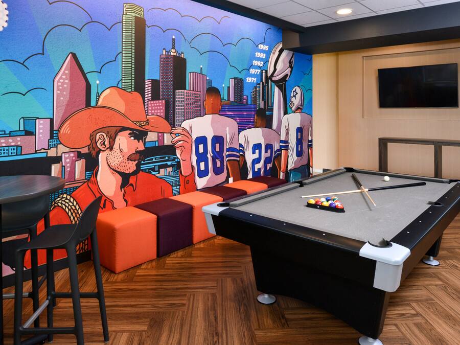 Una mesa de billar con un mural de paisaje urbano, asientos color naranja y un televisor en la pared.
