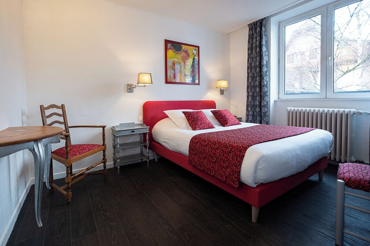 A hotel room with a bed, a chair, a table, a nightstand, a painting, and a window with curtains.