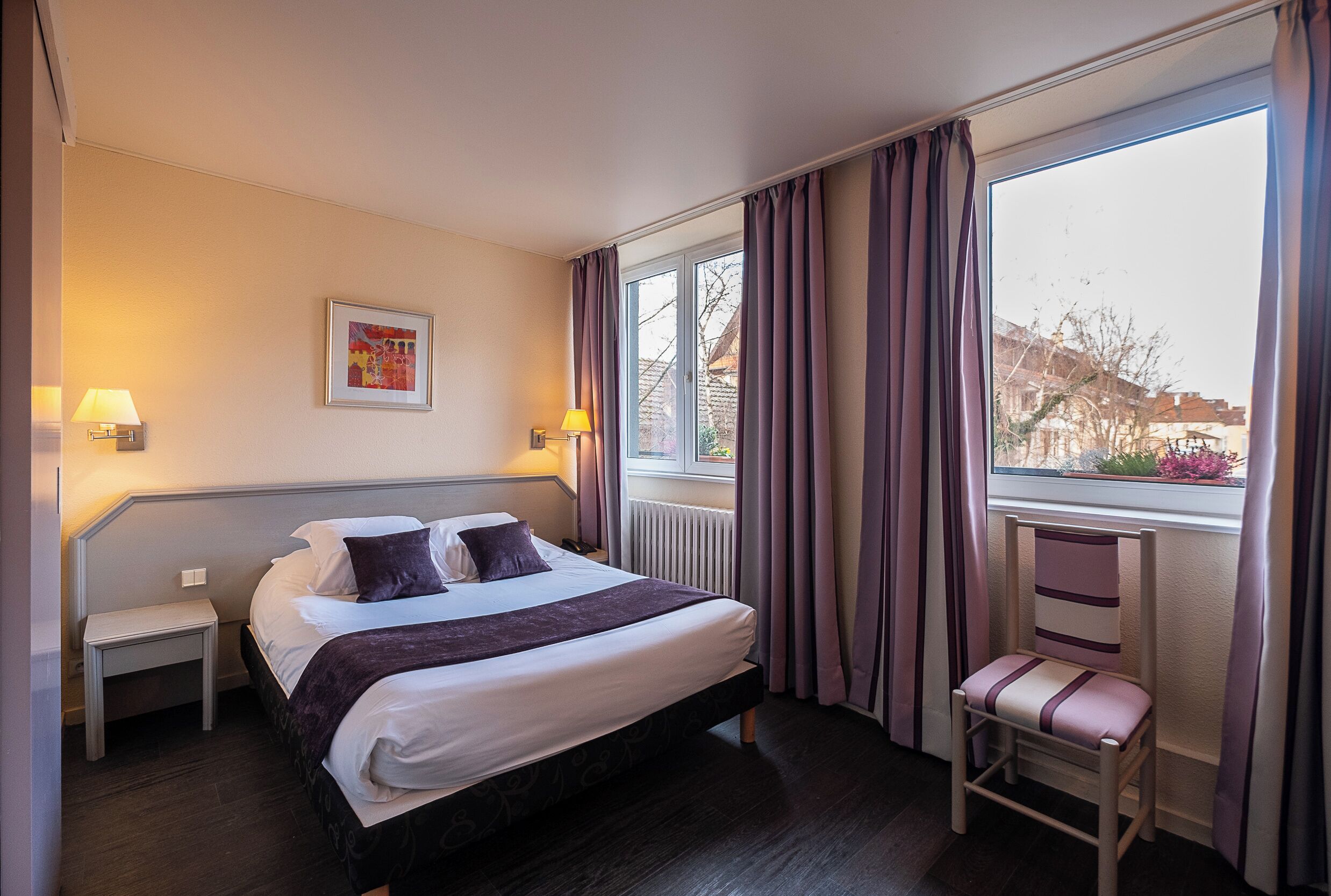 Ein Hotelzimmer mit Bett, Nachttischen, einem Sessel, einem Fenster mit Vorhängen und Blick auf Gebäude.