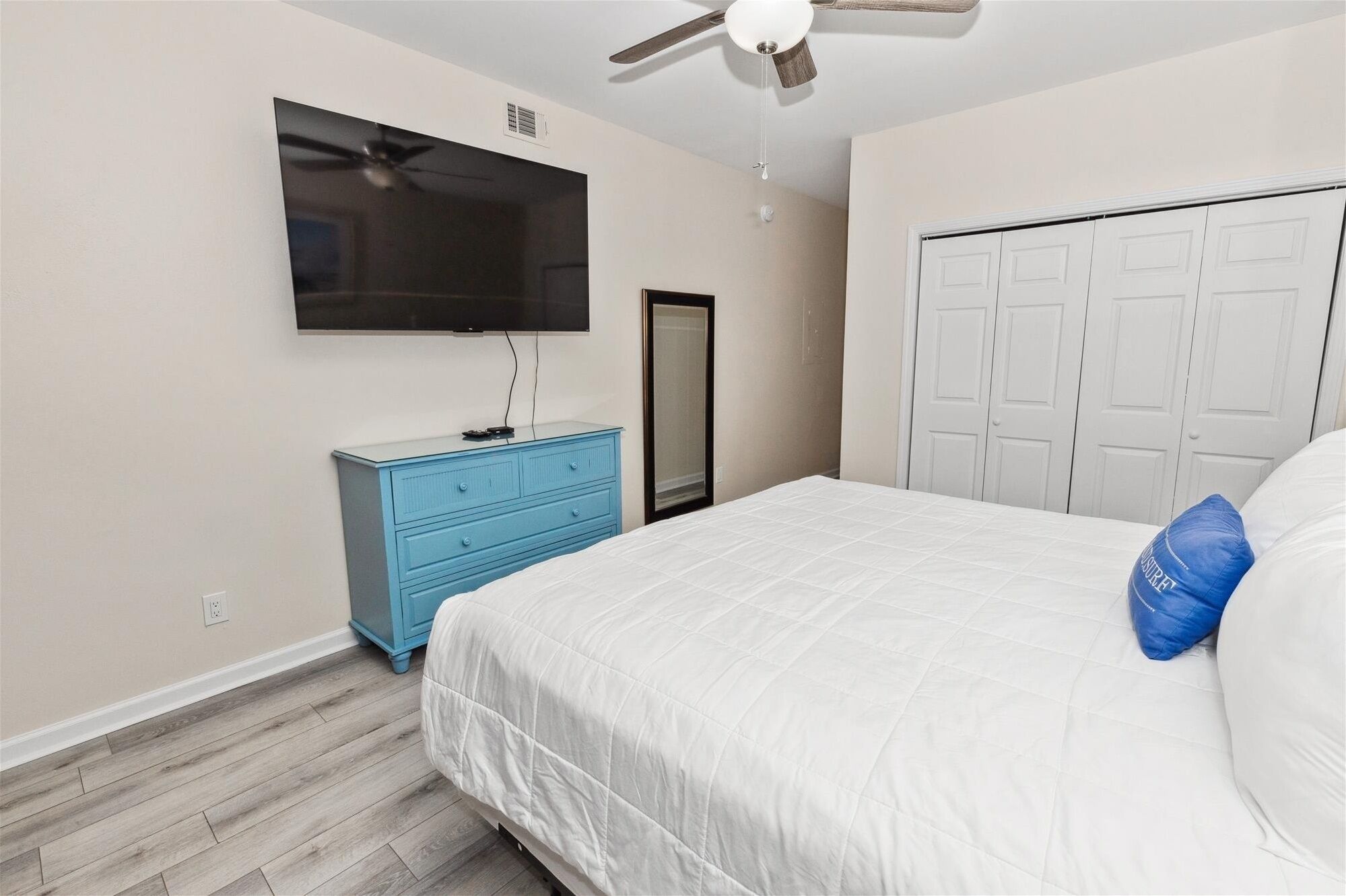 A bedroom with a bed, a blue dresser, a television mounted on the wall, and a ceiling fan.