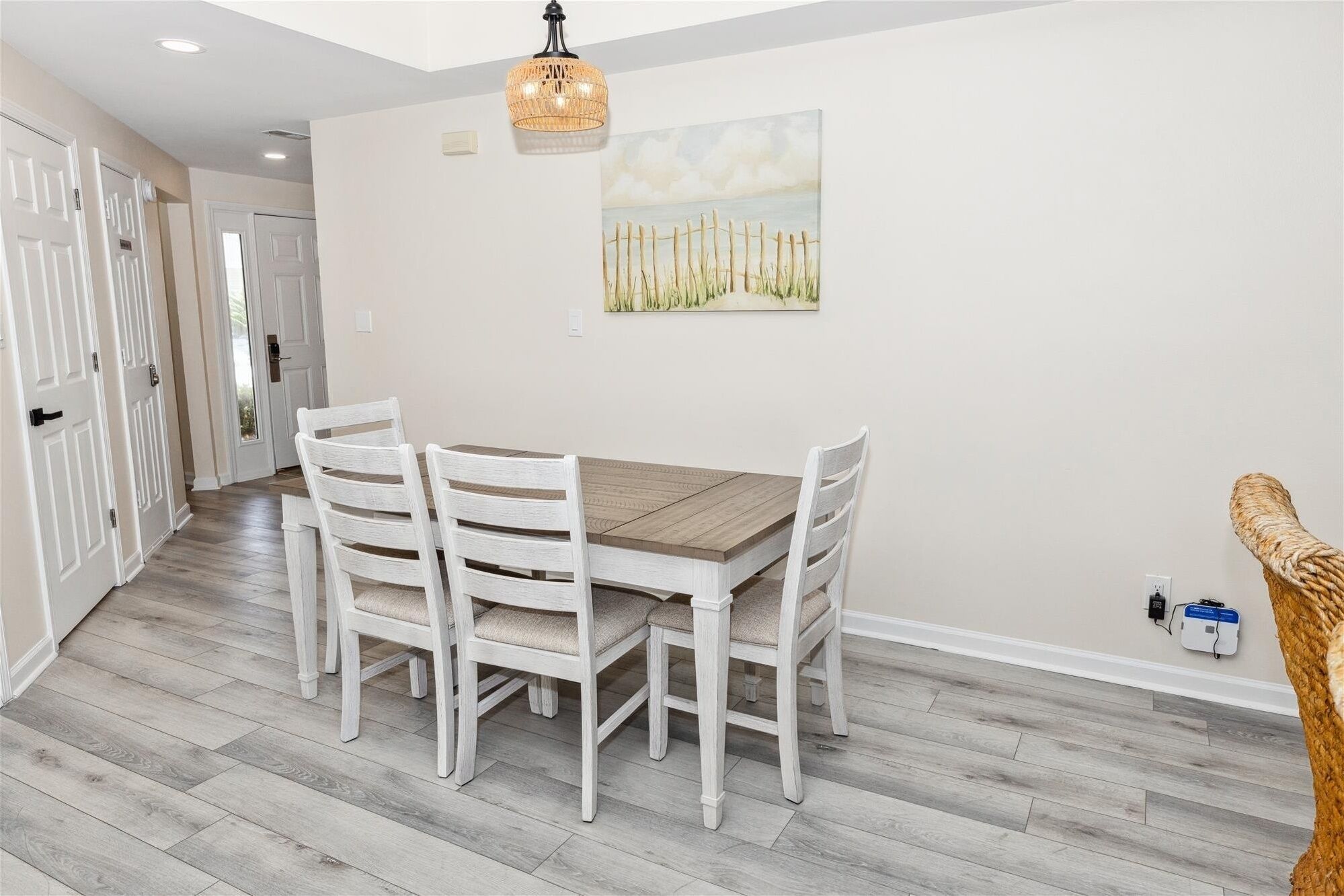 A dining area with a wooden table and white chairs, a hanging light fixture, and a painting on the wall.