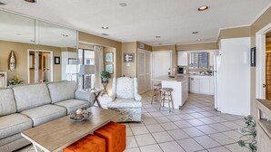 Un salon moderne avec un canapé, une table basse et un coin repas. Au fond, on aperçoit une cuisine équipée d’un réfrigérateur, d’un évier et d’un plan de travail.