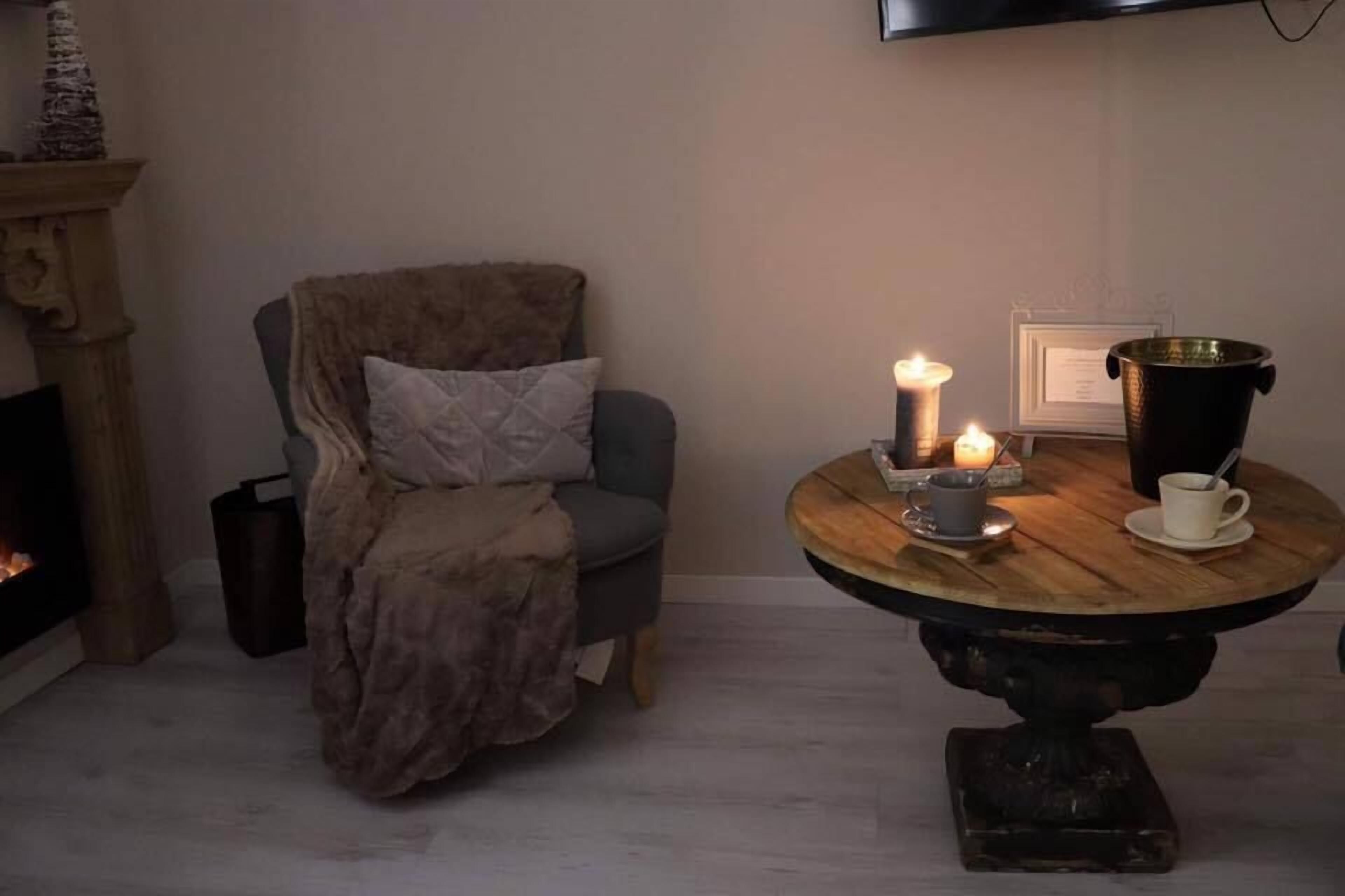A cozy room with a fireplace, a comfortable armchair, a round wooden table with candles, and a brass pot.
