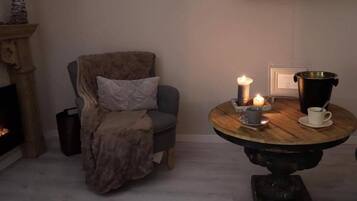 A cozy room with a fireplace, a comfortable armchair, a round wooden table with candles, and a brass pot.