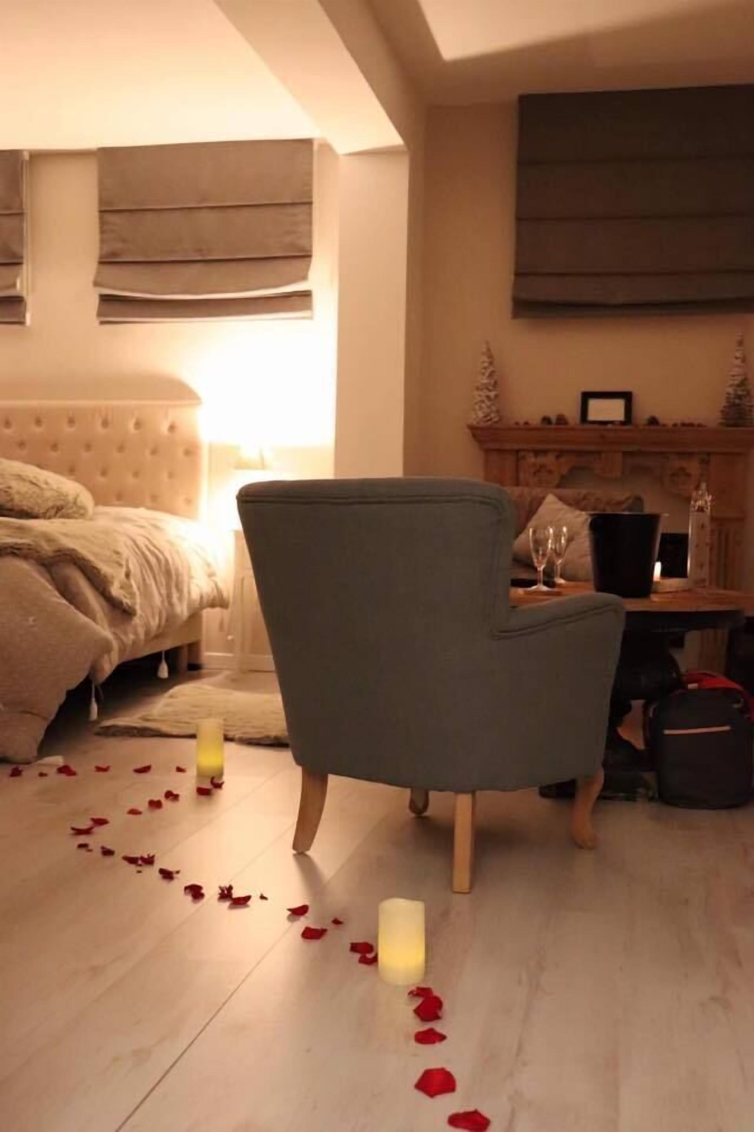 A bedroom with a bed, a chair, a bedside table with a candle, and rose petals on the floor.