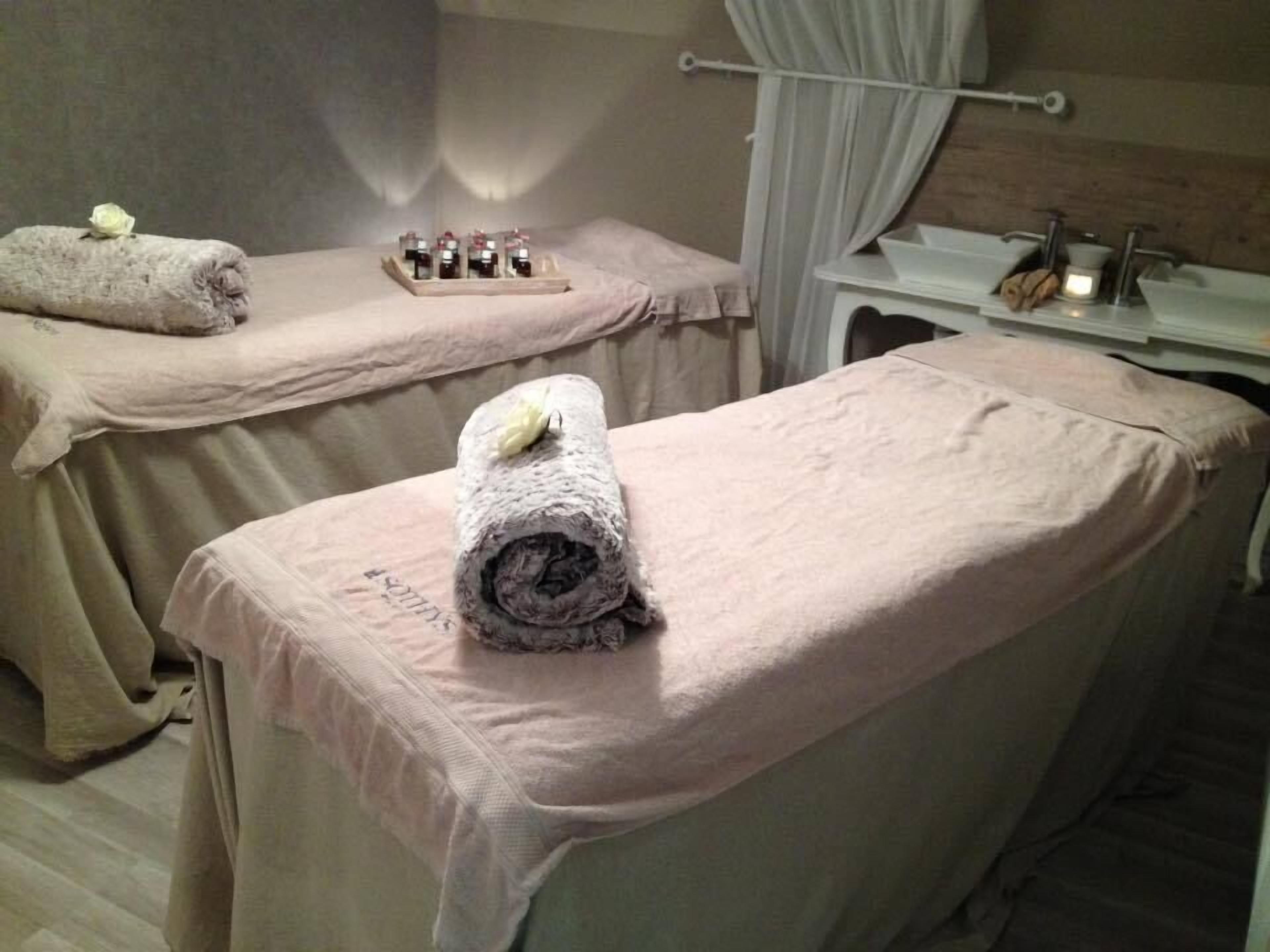 A spa room with two treatment tables covered in beige towels, a shelf with small bottles, and a white sink.