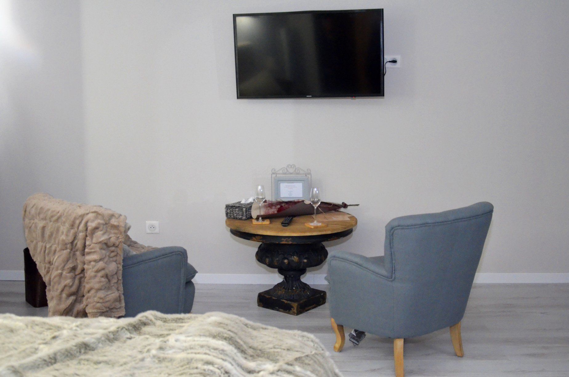 Una habitación moderna con una televisión de pantalla plana montada en la pared, una mesa redonda de madera con una copa de vino y una botella, dos sillones grises y un manto de piel grande sobre la cama.