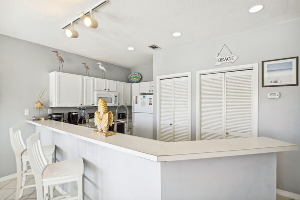 Une cuisine avec des meubles blancs, un îlot central blanc doté de tabourets, un réfrigérateur, une horloge et un panneau décoratif représentant une plage accroché au mur.