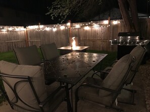 Een terras buiten met een metalen tafel en vier stoelen, een barbecue en lichtjes aan een houten schutting.