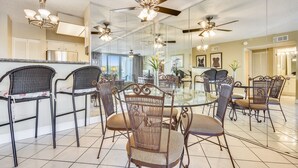 A dining area with a glass table surrounded by wicker chairs, ceiling fans, and a kitchen in the background.