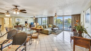 A spacious living room with a large sofa, dining table, and chairs, a balcony with a view, and ceiling fans.