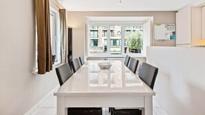 A modern dining area with a long table and chairs, large windows with curtains, and a view of a residential building. - Campanula (Koksijde)