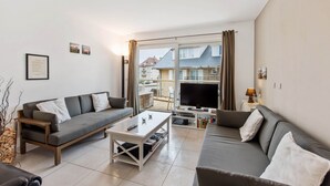 A modern living room with a sofa, coffee table, TV, and a view of a residential area through large windows. - Campanula (Koksijde)
