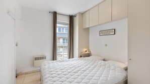 A bedroom with a large bed, a window with curtains, a bedside table with a lamp, and a wall-mounted sign. - Campanula (Koksijde)