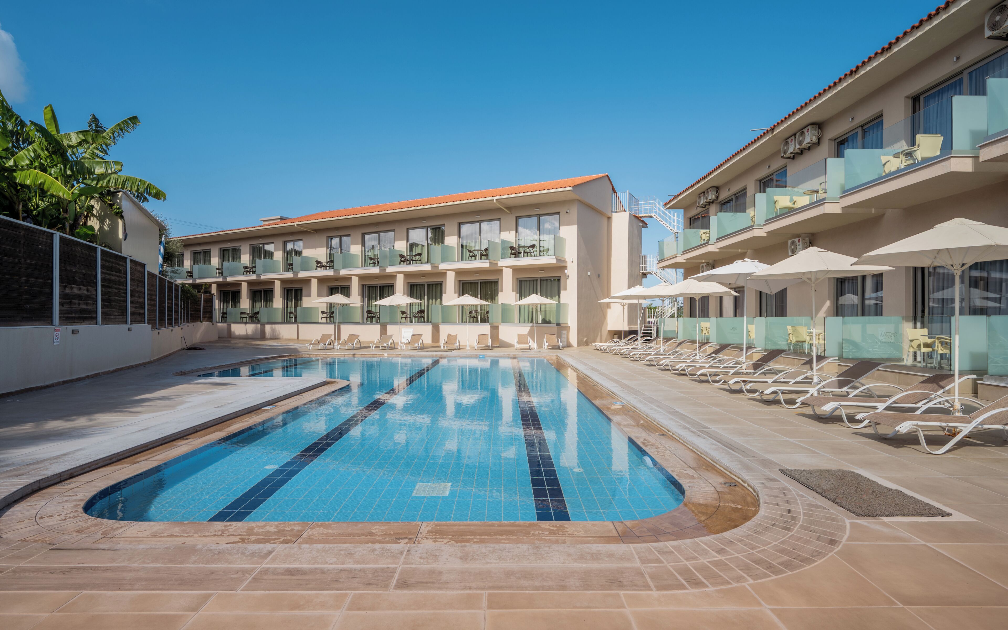 Una piscina con tumbonas y sombrillas, un edificio con balcones y un cielo azul despejado.