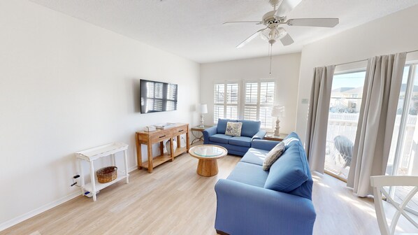 Ein modernes Wohnzimmer mit einem blauen Sofa, einem Holztisch, einem weißen Deckenventilator und großen Fenstern mit Jalousien.