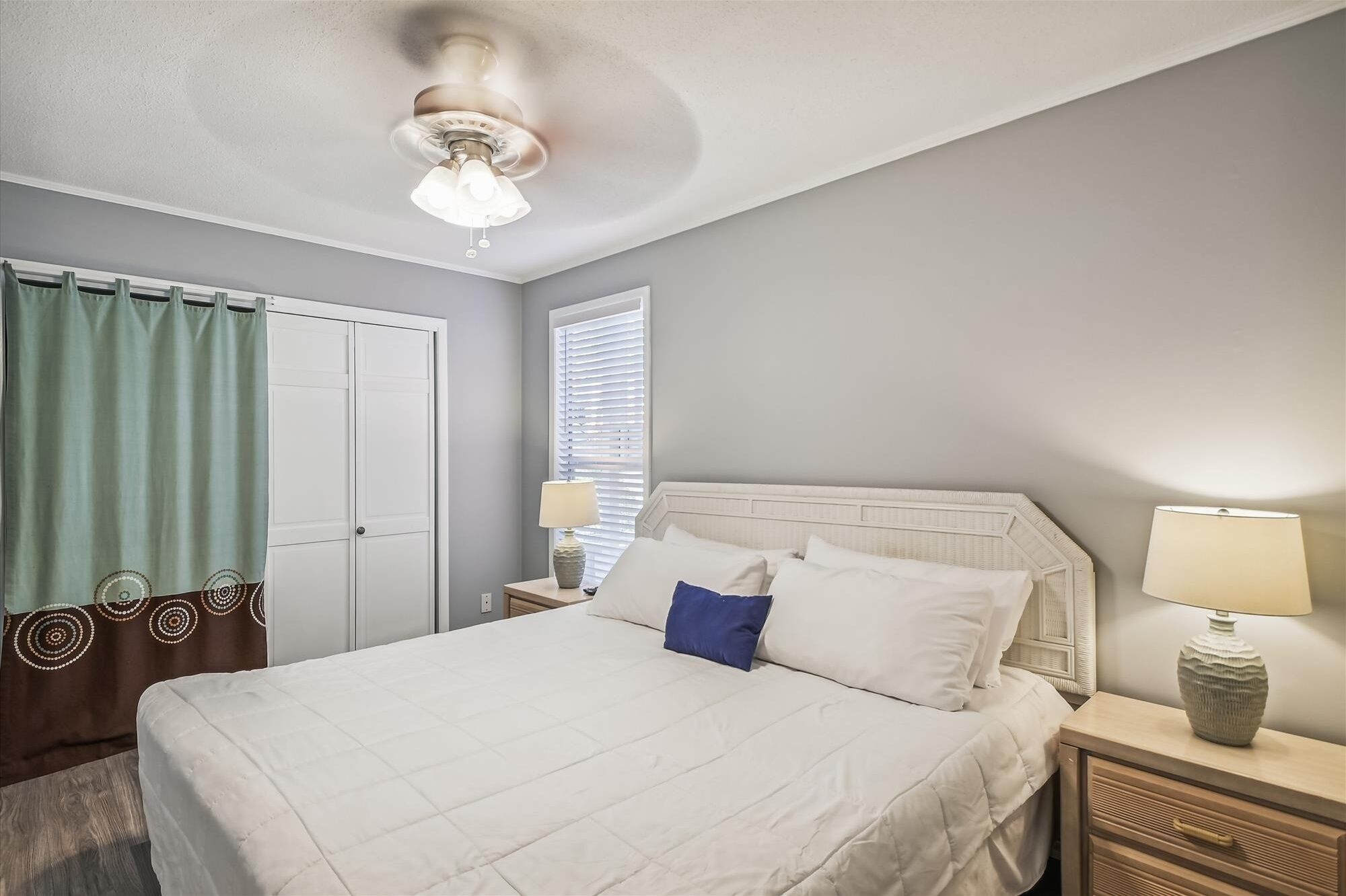 Une chambre d’hôtel comprenant un lit, des tables de chevet, une lampe, une fenêtre avec des stores, un ventilateur de plafond et une porte avec un rideau.