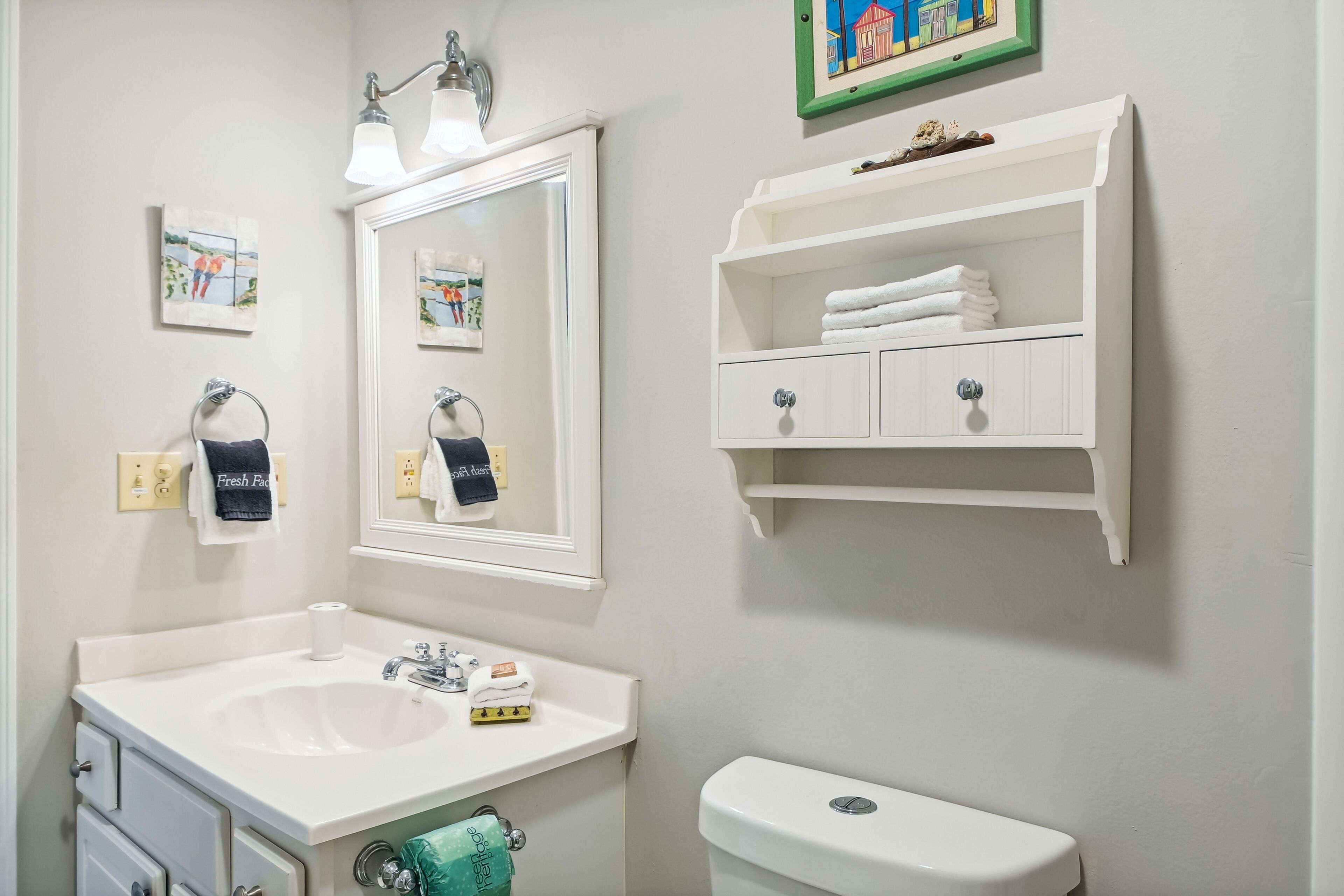 A bathroom with a white sink, mirror, and toilet. There are towels and a soap dispenser on the sink, and a shelf with towels above the toilet.