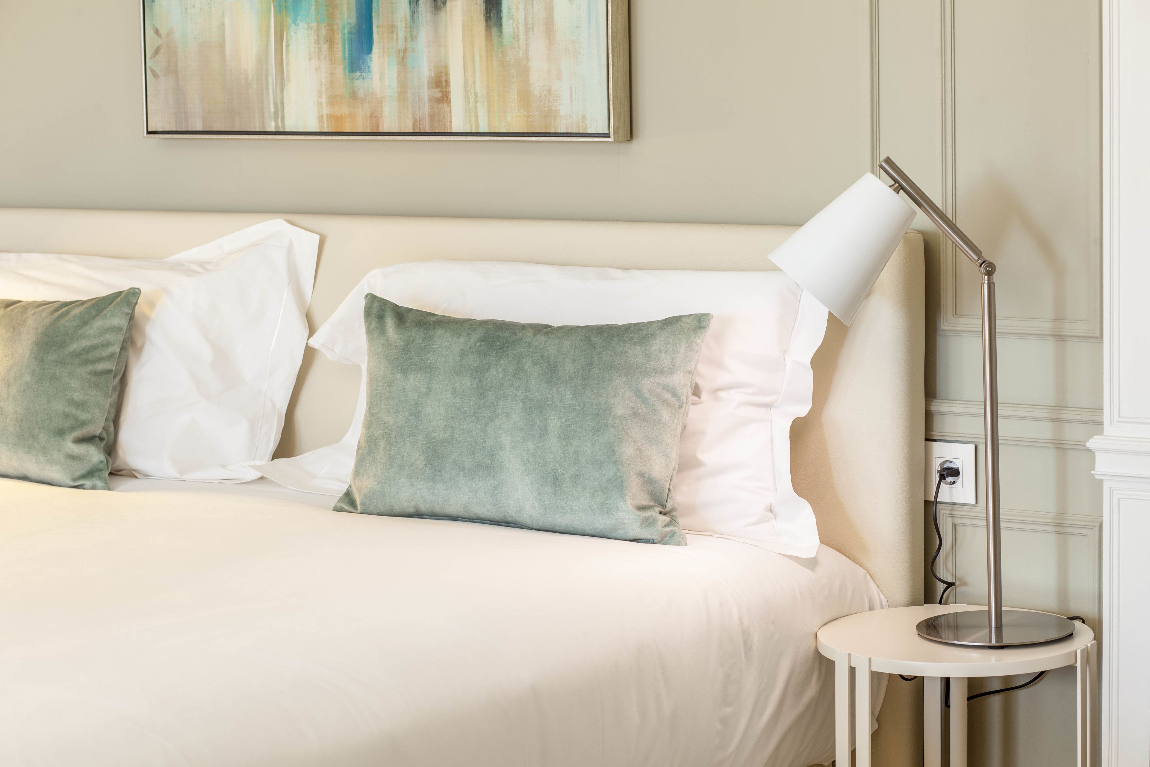 Un lit bien fait, avec des oreillers blancs et verts, une table de chevet avec une lampe, et un tableau accroché au mur.